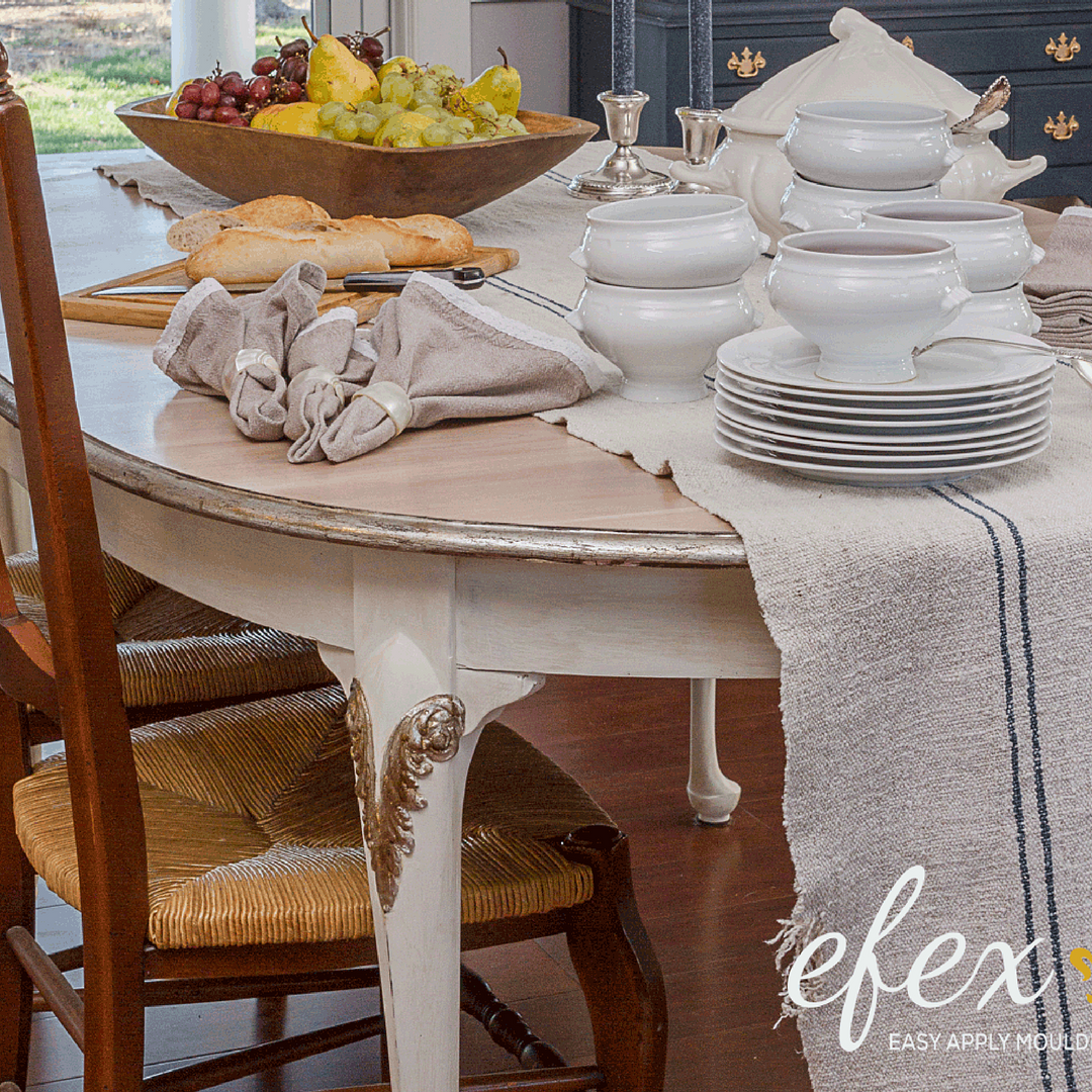 Cream painted oval dining table with a gilded L1 Small Queen Anne Leg Efex flexible moulding on the leg, set with white dishes, linen napkins, bread and fruit