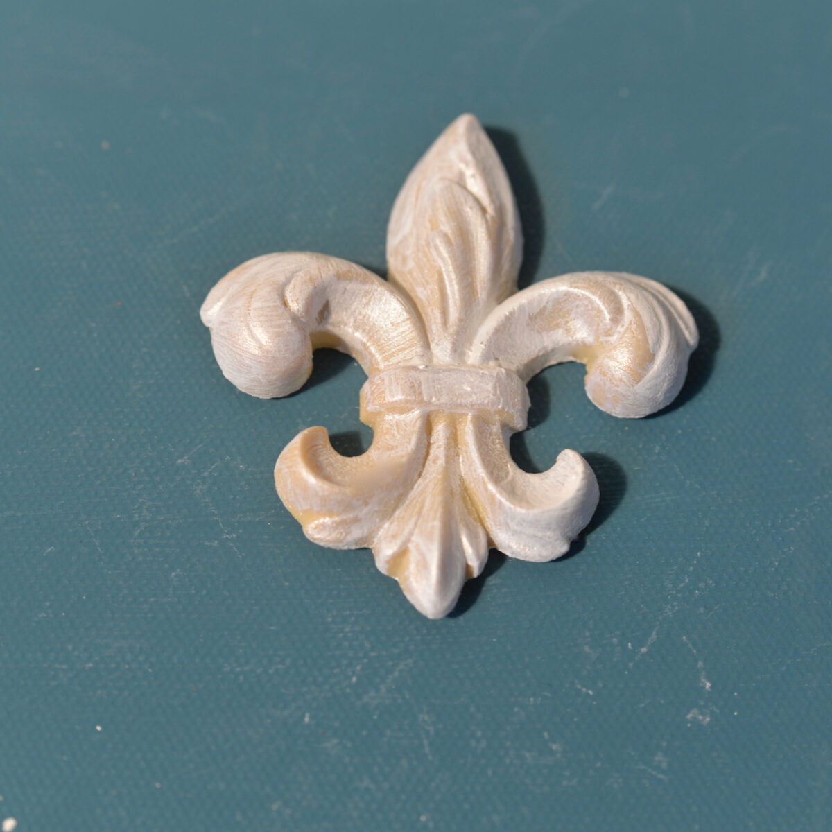 FDL3 Fleur de Lis Efex flexible moulding applique with a pearlescent painted finish, on a teal surface