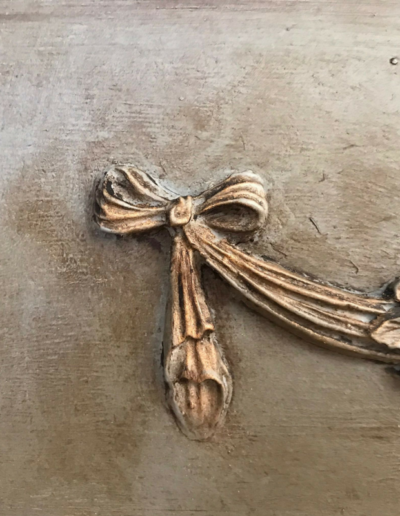 Close-up of the ribbon bow end of an SW9 Rose Swag Efex flexible moulding, antiqued in gold on a distressed painted surface