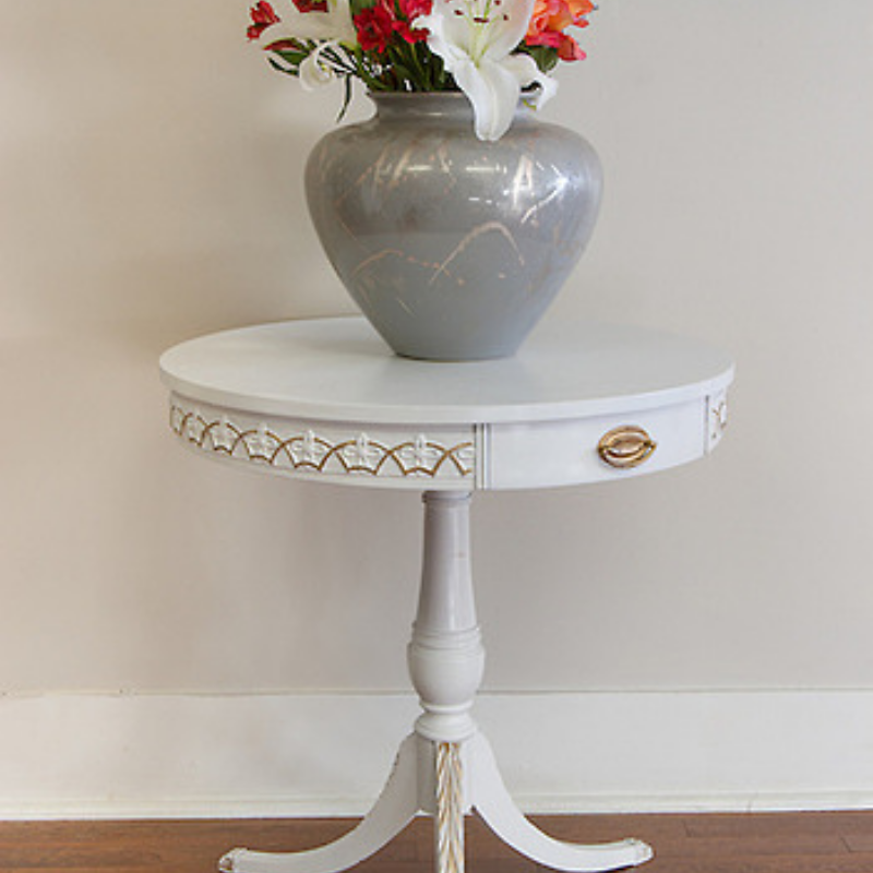White pedestal drum table with gold-accented T23 Celtic Art Deco Efex flexible moulding around the apron, topped with a grey vase of lilies
