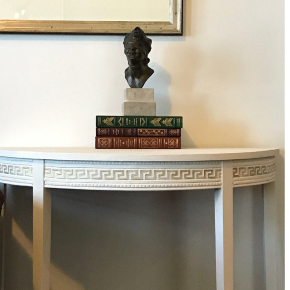White demilune console table with T33 Greek Key Trim Efex flexible moulding running around the curved apron, styled with books and a bronze bust